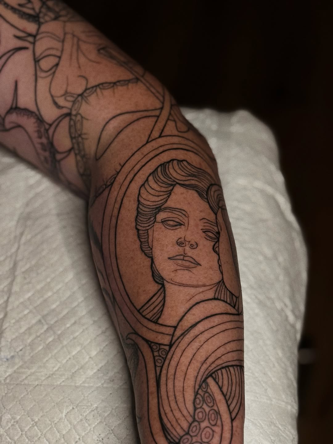 Portrait of a person with wavy hair in black and grey ink on forearm, surrounded by decorative swirling linework and ornamental details.