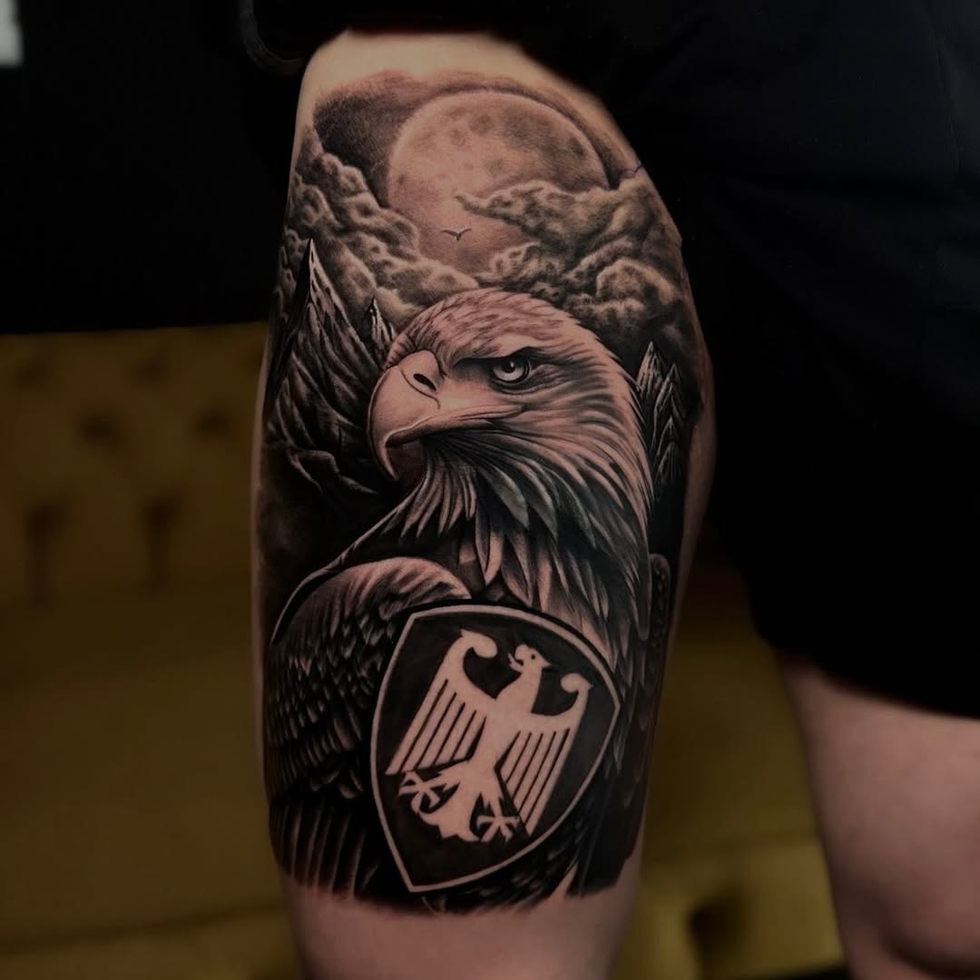 Bald eagle head with detailed feathers and intense gaze above a German coat of arms shield on upper arm, black and grey shading.