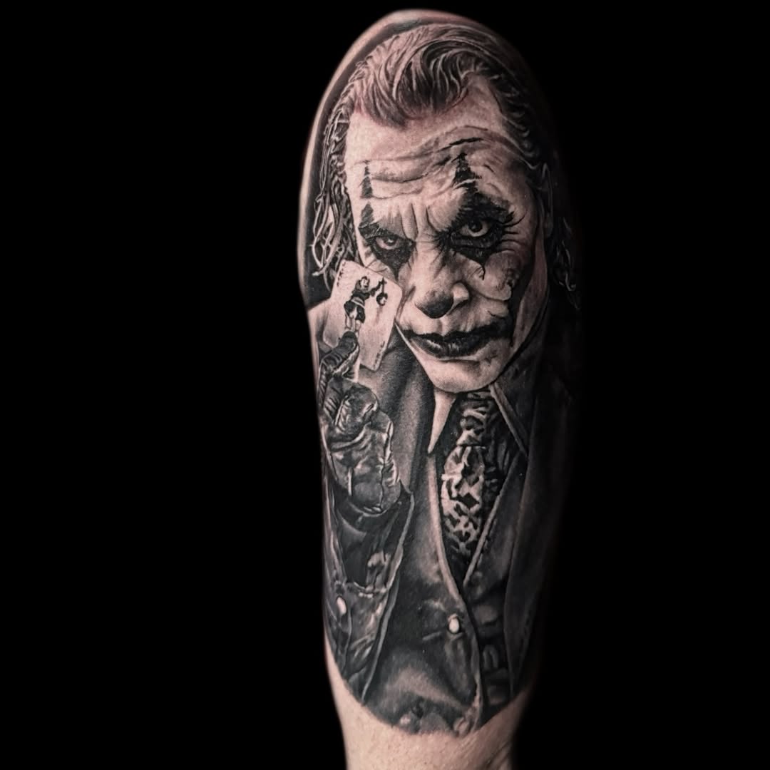 A black and grey portrait tattoo on an upper arm depicting a character with theatrical makeup, dramatic facial expressions, and formal attire with intricate shading details.
