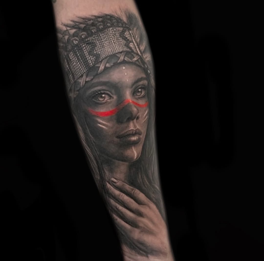 Portrait of a woman with long hair wearing a mesh headpiece and red eye mask, tattooed on forearm in black and grey with smooth shading.