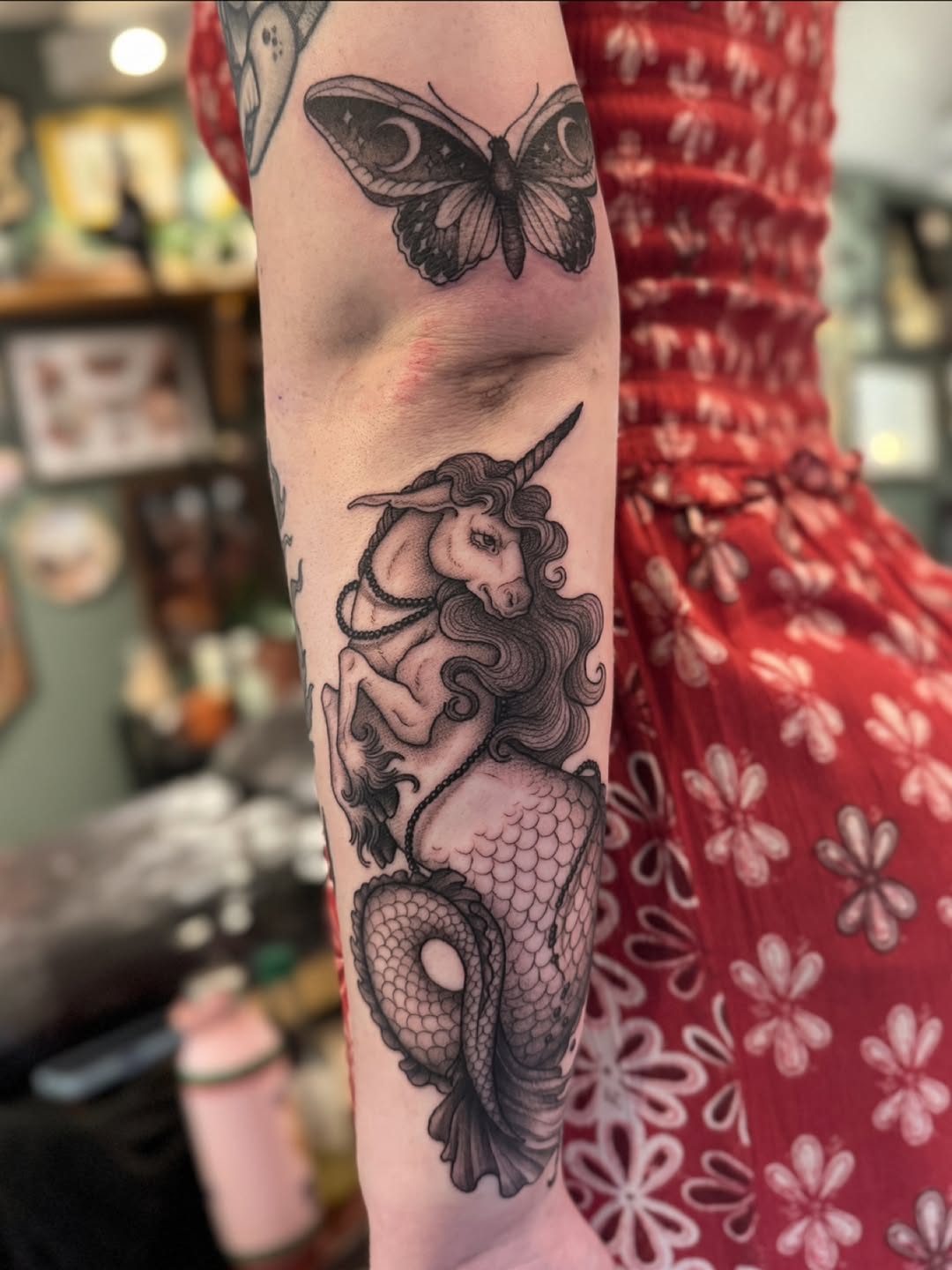 Arm tattoo featuring a unicorn mermaid with flowing hair and scaled tail, accompanied by a detailed moth above, rendered in black and grey with soft shading.