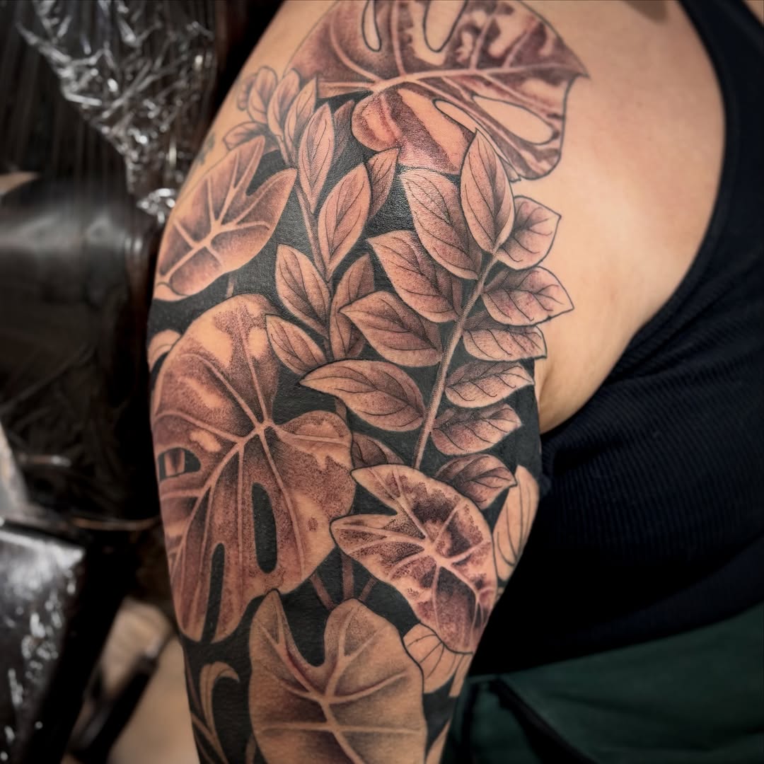Botanical sleeve tattoo featuring layered leaves and flowers with soft shading in black and grey on upper arm.