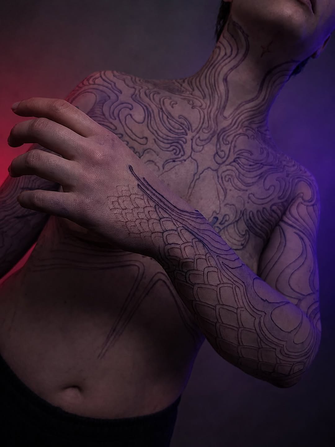Black and grey ornamental tattoo covering chest and arm with flowing baroque-style scrollwork and scale pattern details.