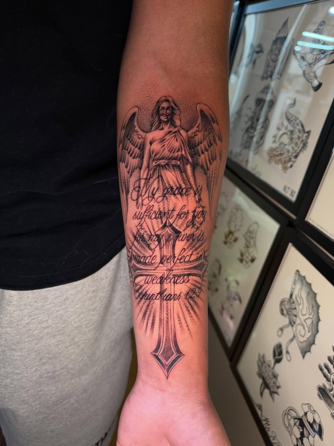 Winged angel figure with flowing robes and script text on forearm, rendered in black and grey with soft shading and a cross below.