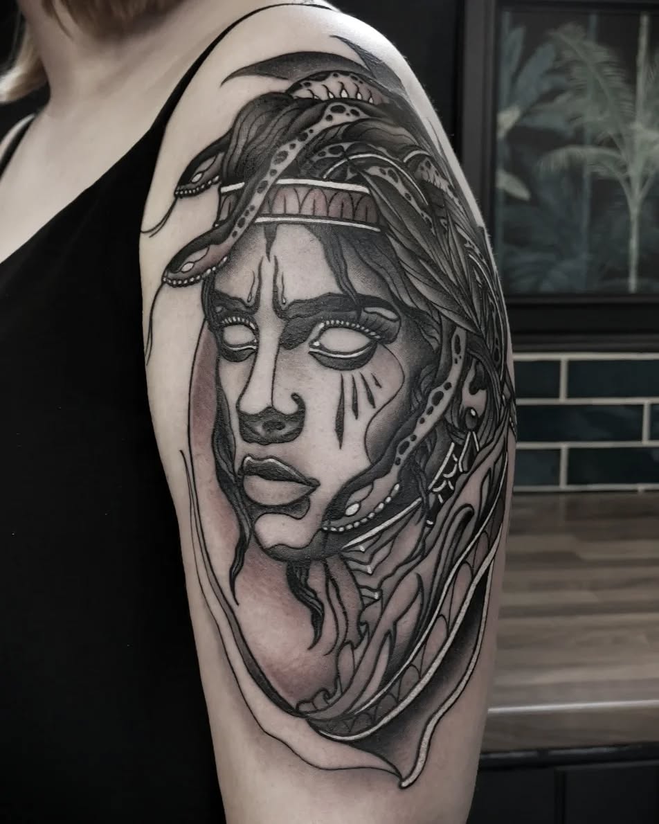 Black and grey portrait tattoo of a woman with closed eyes, ornate headdress, and decorative neck jewelry on upper arm.