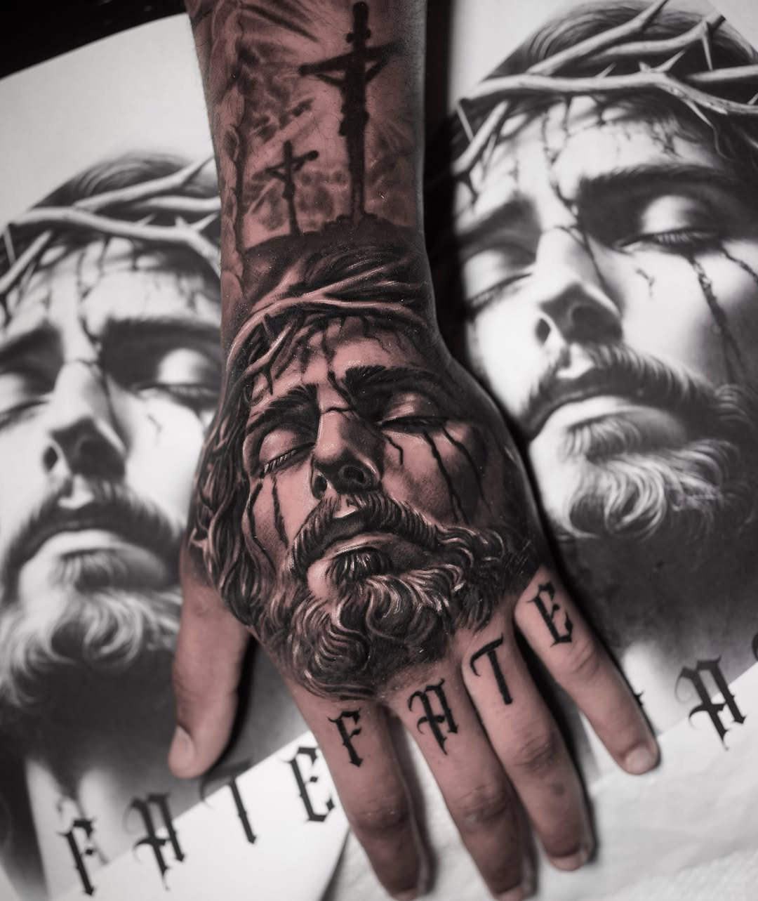 Black and grey hand tattoo of Jesus Christ's face wearing a crown of thorns, with crucifixion scene on wrist and 'FATE' lettering on fingers.