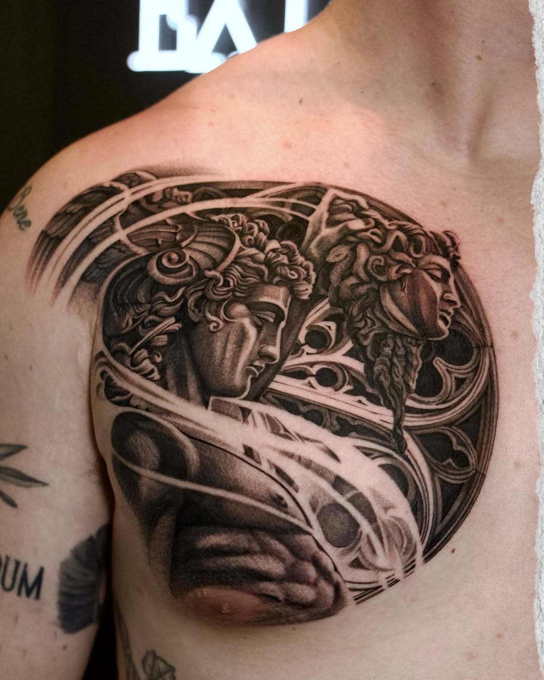 Classical sculptural relief tattoo featuring two profile heads with ornate decorative elements on upper arm, rendered in black and grey with smooth shading.