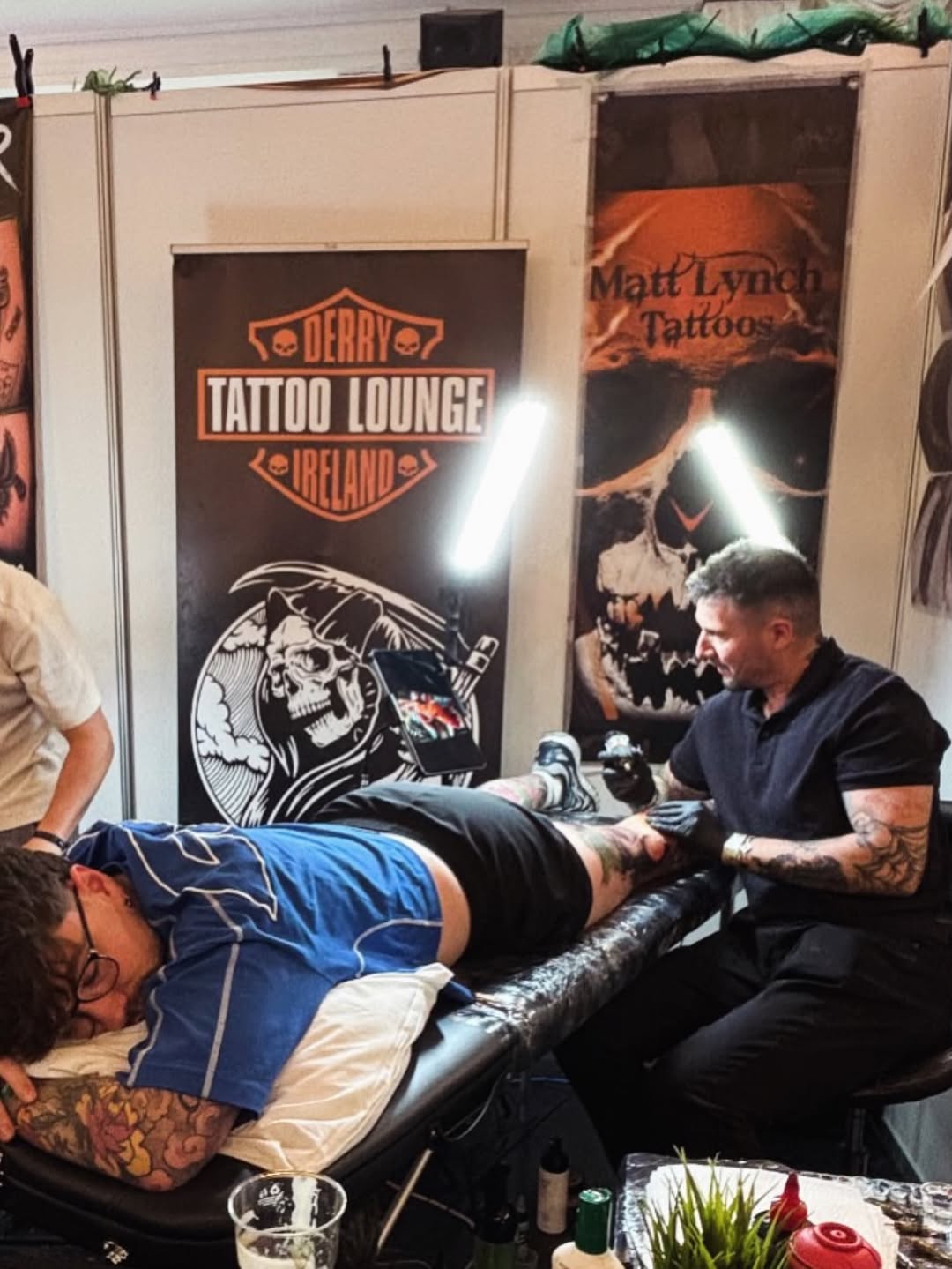 Tattoo artist applies black and grey ink to client's leg, creating a detailed design with smooth shading and gradients.