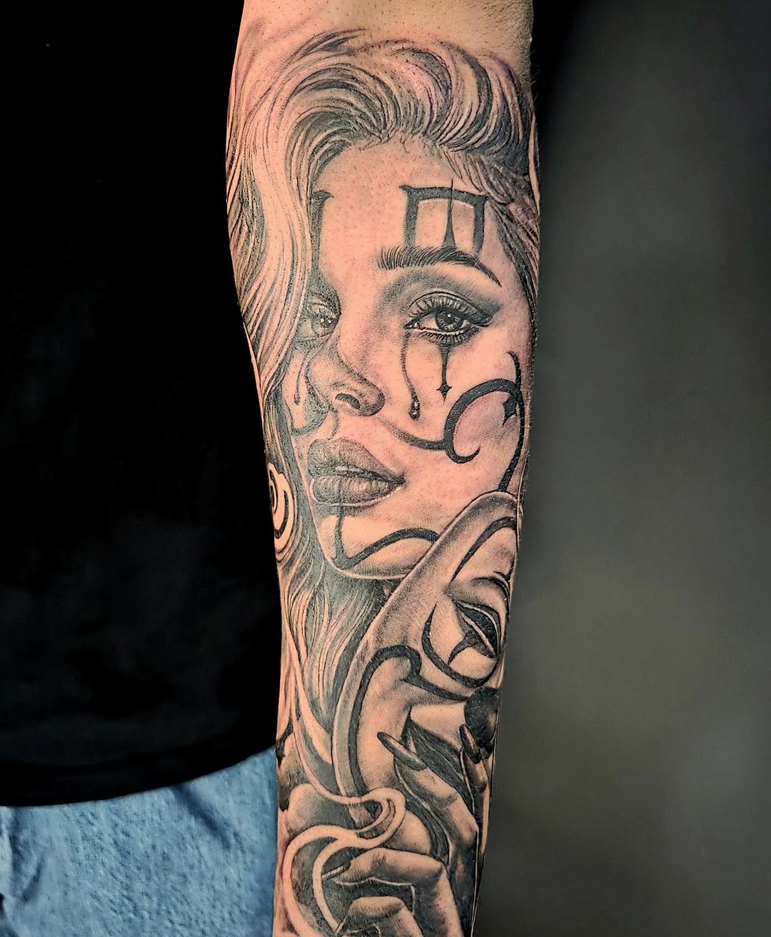 Portrait tattoo of a woman with wavy hair, tears, geometric forehead design, and ornamental neck flourishes on forearm in black and grey.