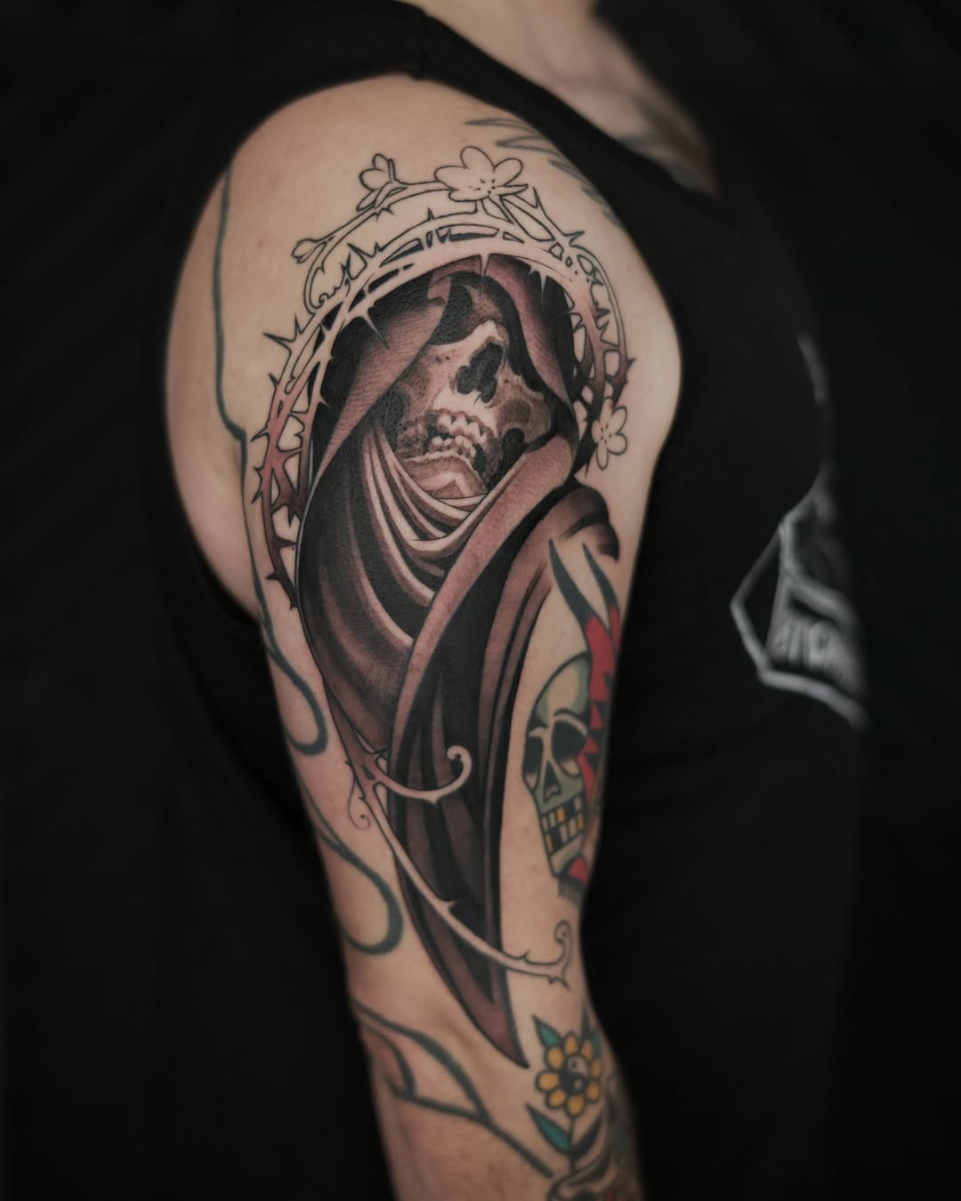 Hooded skeleton figure with ornate floral frame on upper arm, rendered in black and grey with soft shading.