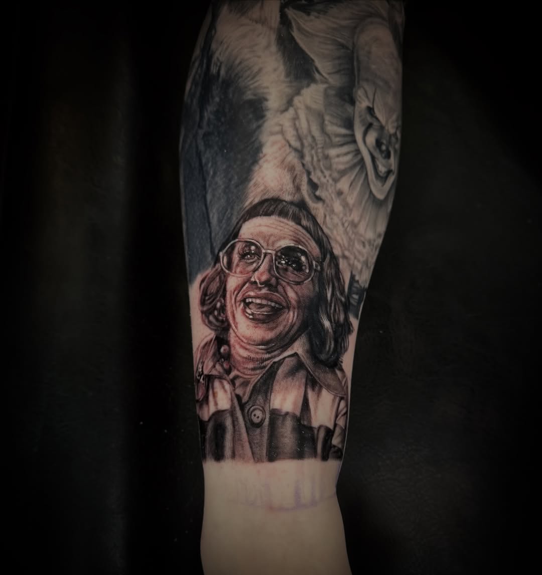 Forearm tattoo featuring a smiling woman with glasses and curly hair, above an owl and a menacing clown face, black and grey shading.