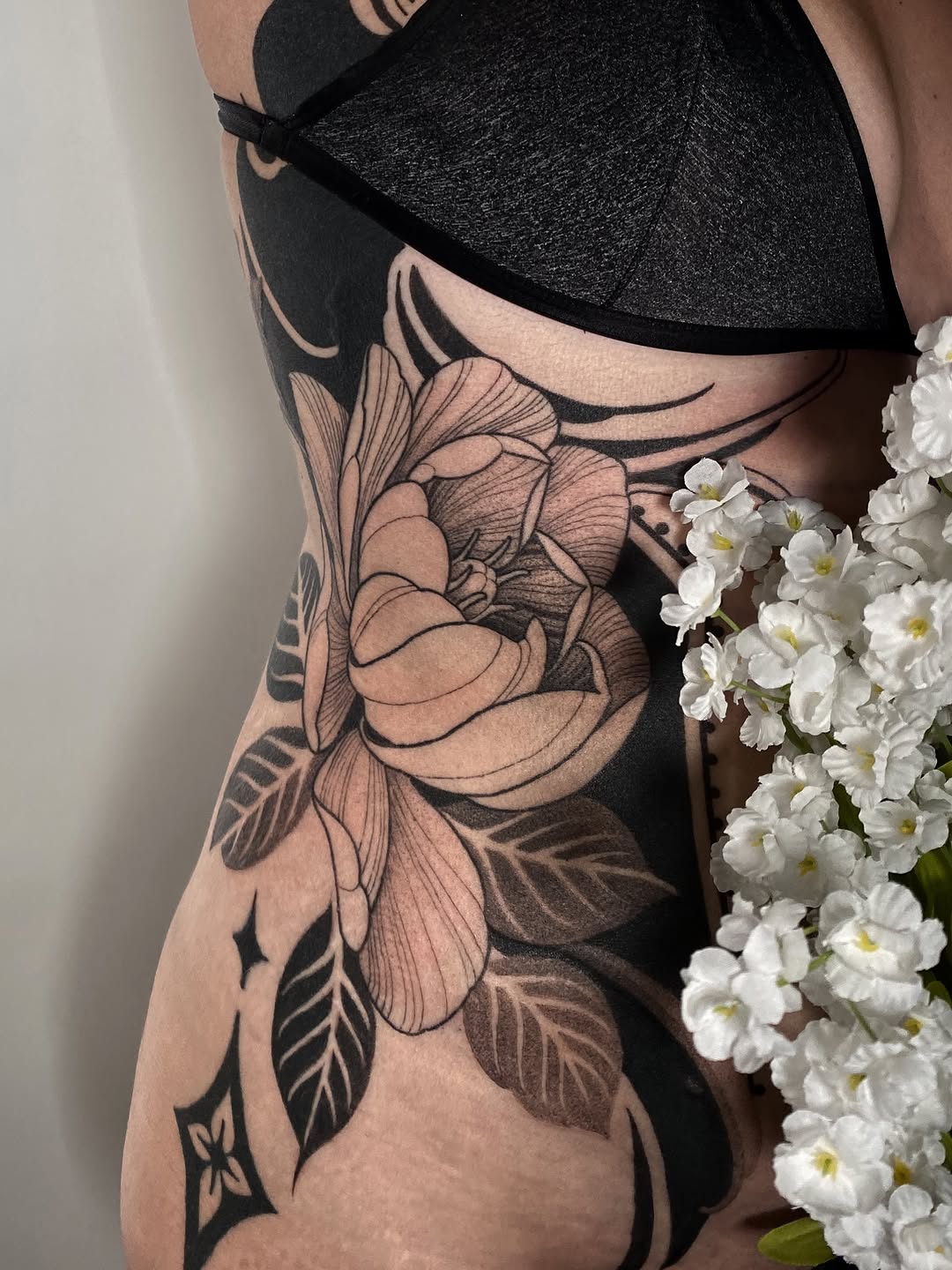 Large floral tattoo with detailed peony and leaves on ribcage in black and grey with smooth shading.