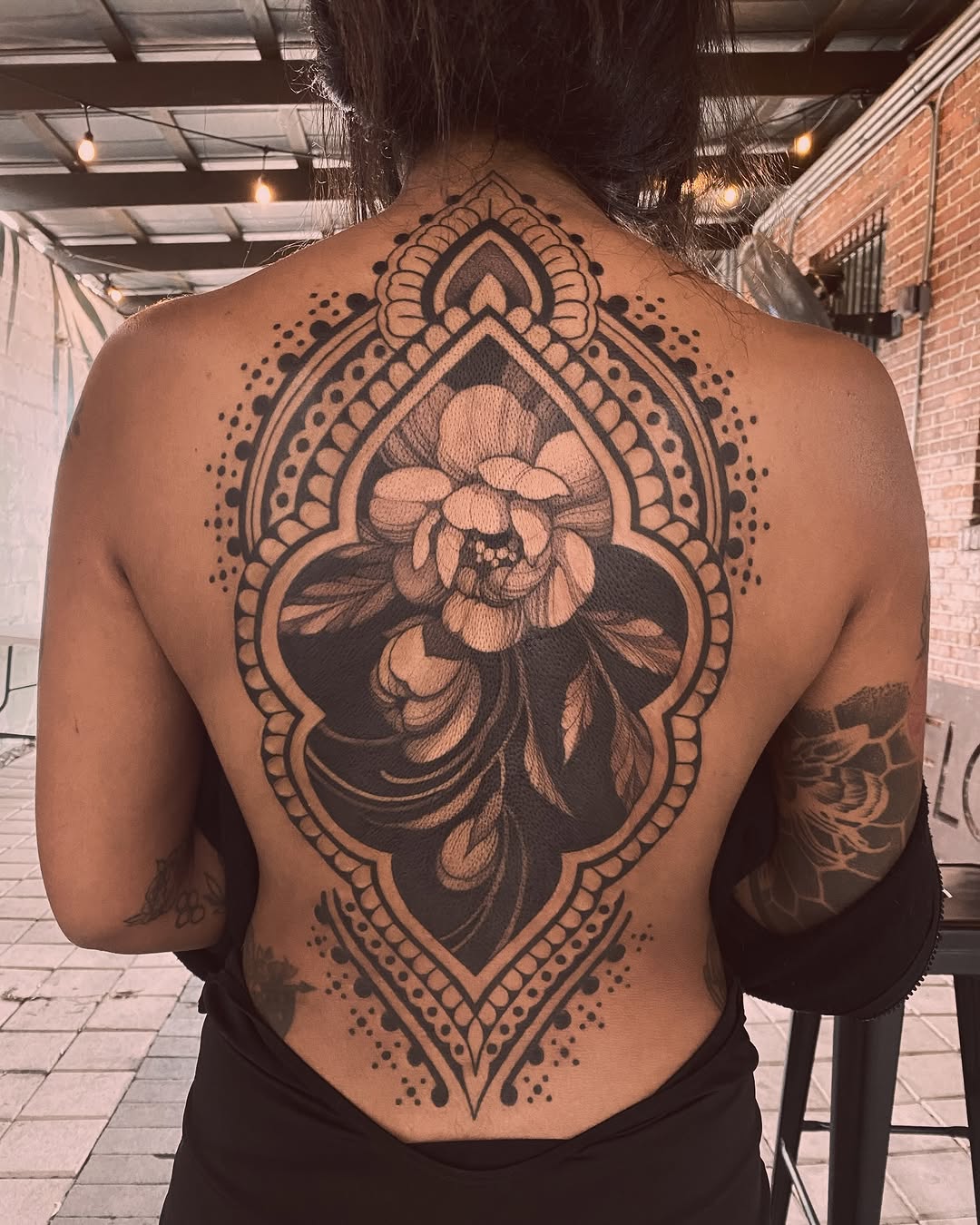 Large black and grey mandala-framed peony flower tattoo covering the upper back and spine with ornate decorative borders.