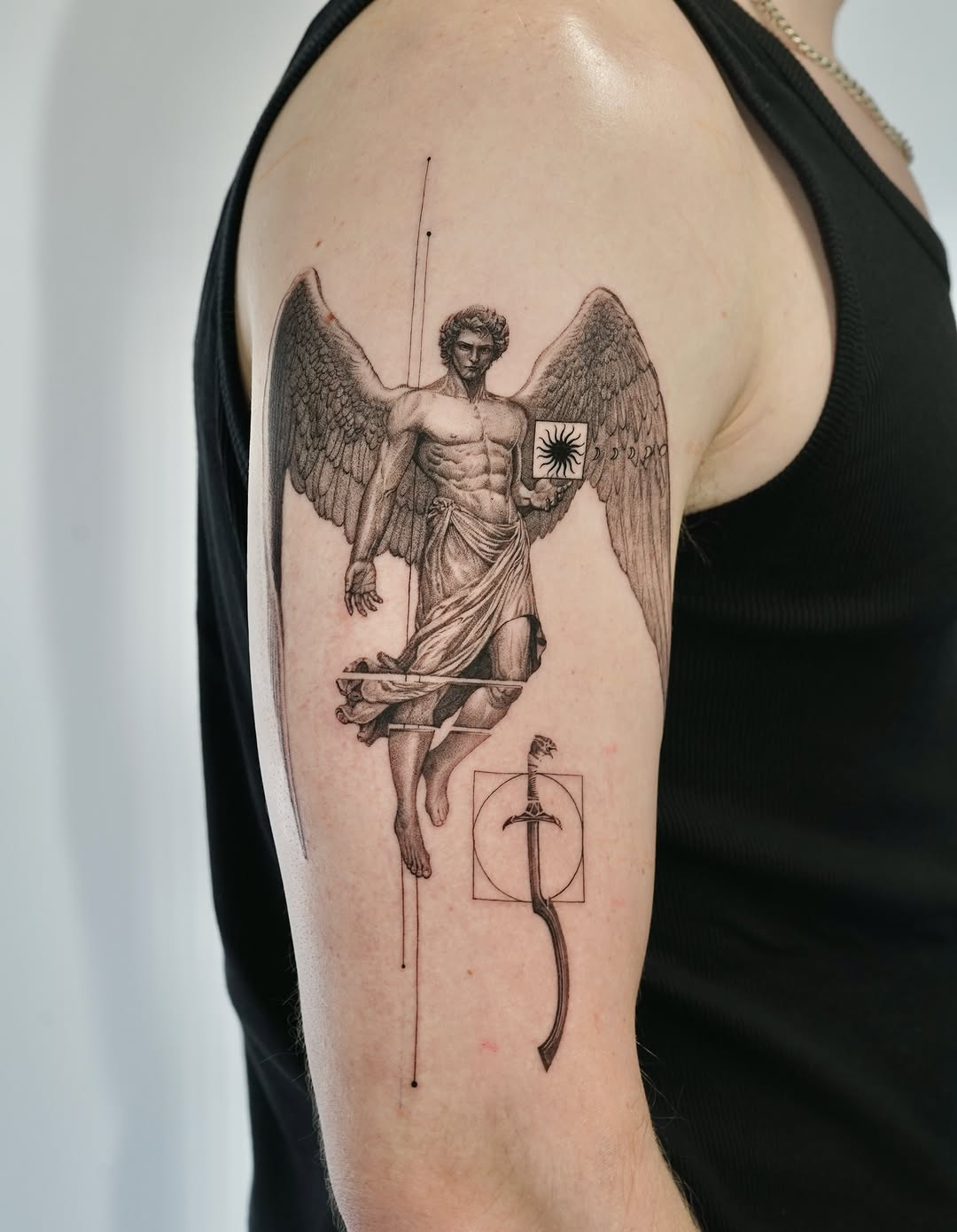 Winged angelic figure with muscular torso holding a sword and sun symbol, adorned with geometric elements, tattooed on upper arm in black and grey.