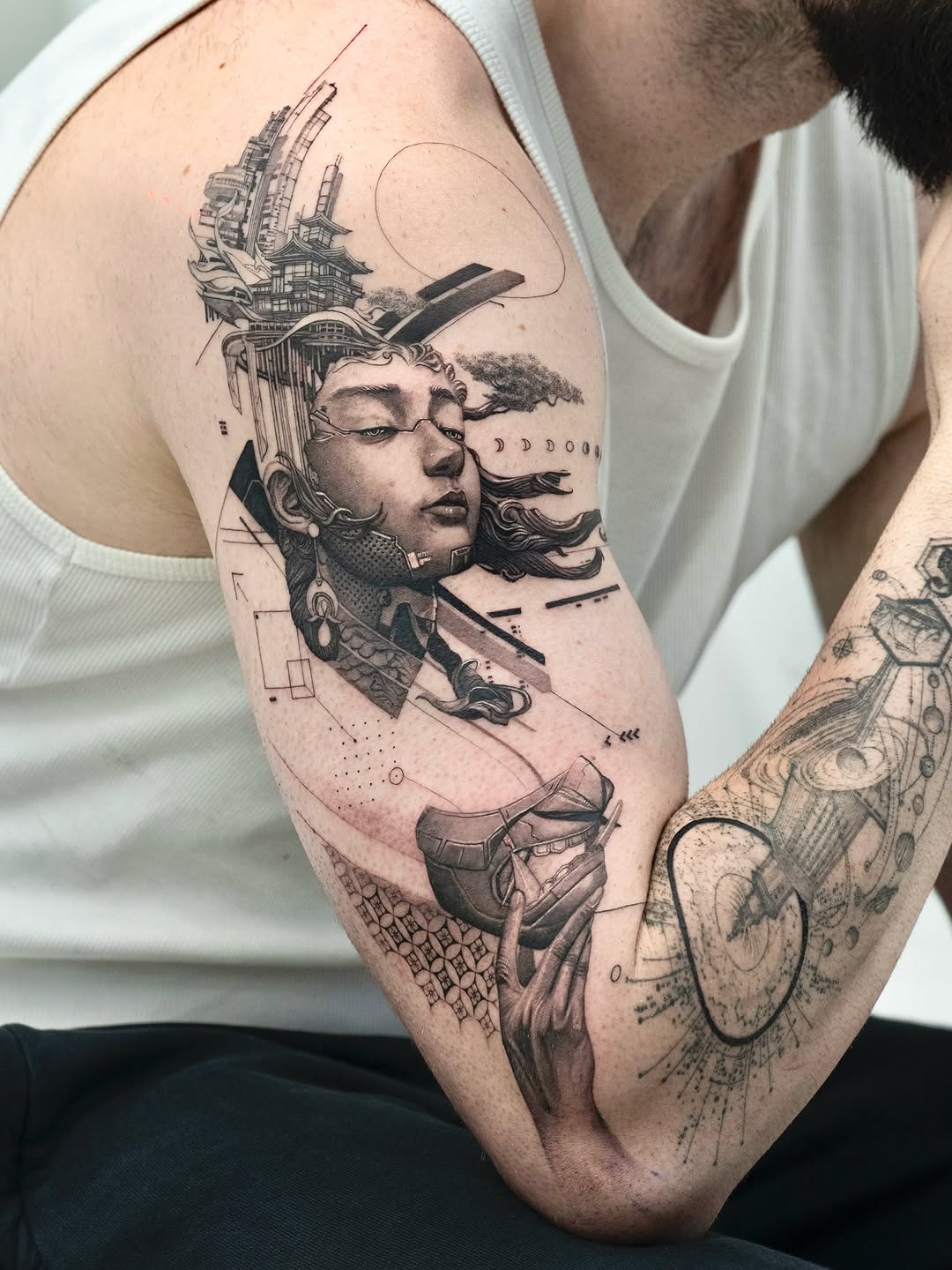 Black and grey portrait tattoo on upper arm featuring a woman's face with architectural buildings emerging from her head, geometric line work, and decorative elements.