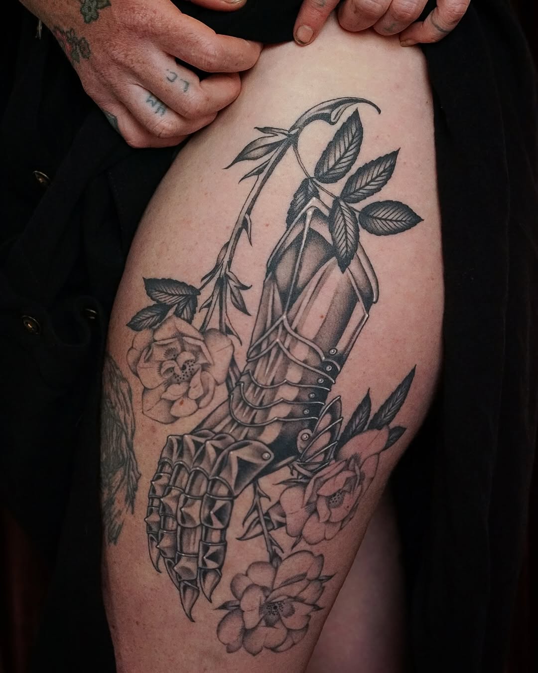 Anatomical skeletal hand with ornate armor plating and botanical vines featuring roses, rendered in black and grey shading on thigh.