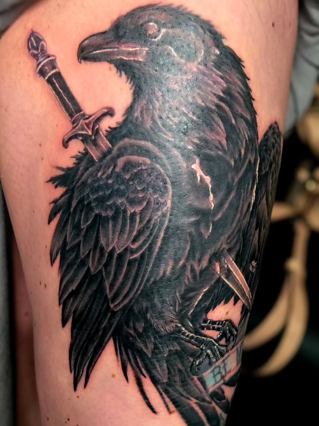 Black and grey raven tattoo on upper arm holding a dagger and banner, rendered with detailed feather shading.