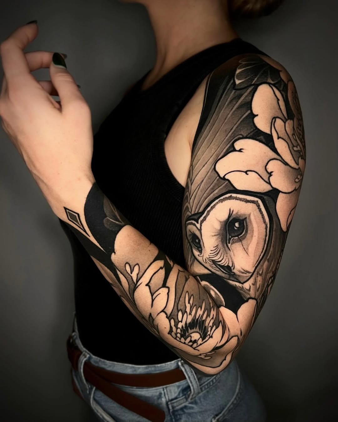 Full arm sleeve tattoo featuring an owl with spread wings, skull, and botanical elements in black and grey shading on person's right arm.