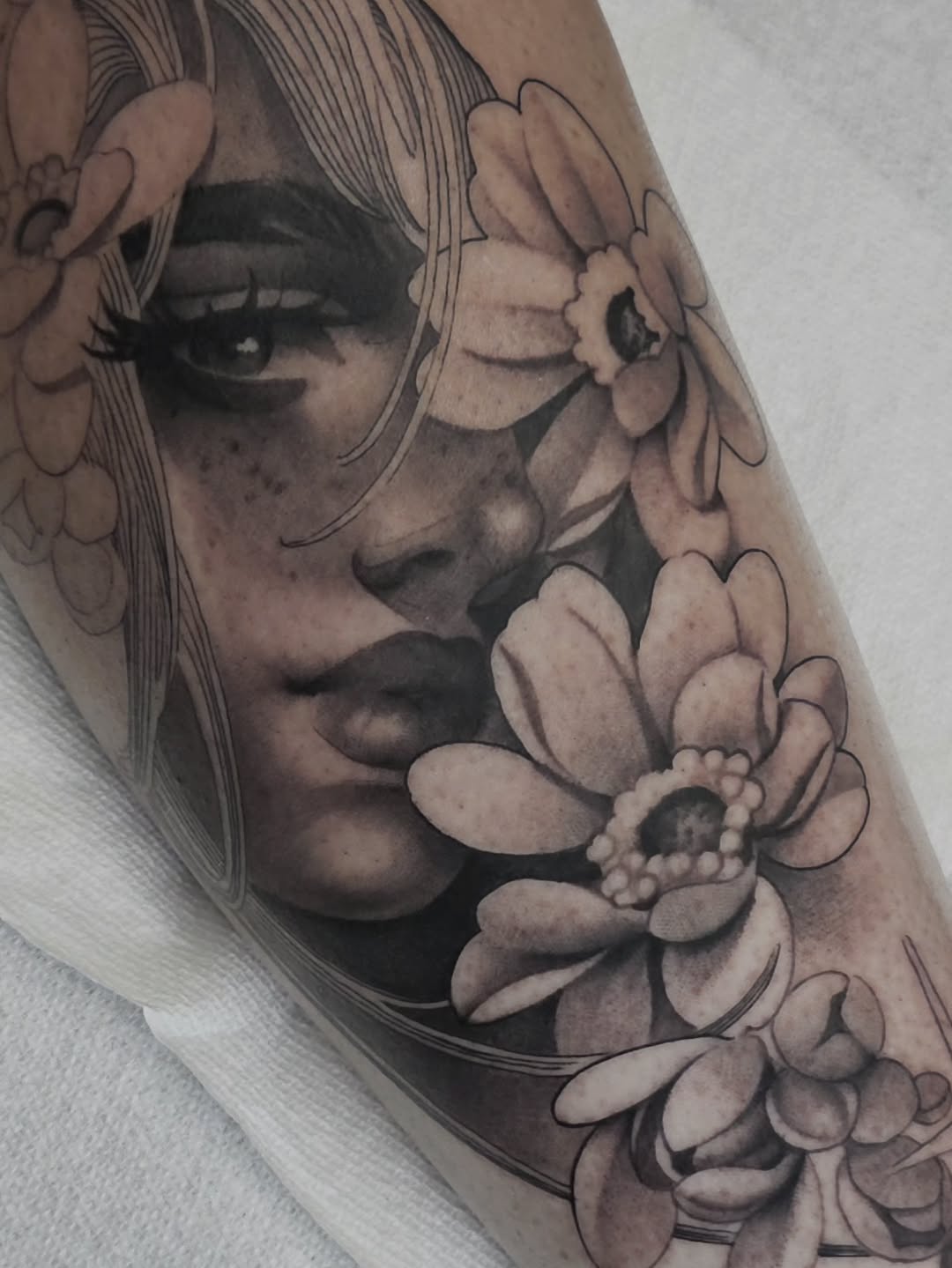 Black and grey portrait tattoo of a woman's face with flowing hair and large flowers on the forearm, rendered in soft shading.