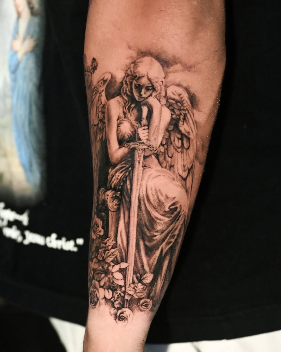 Winged angelic female figure in flowing robes surrounded by roses on forearm, rendered in black and grey with soft shading.
