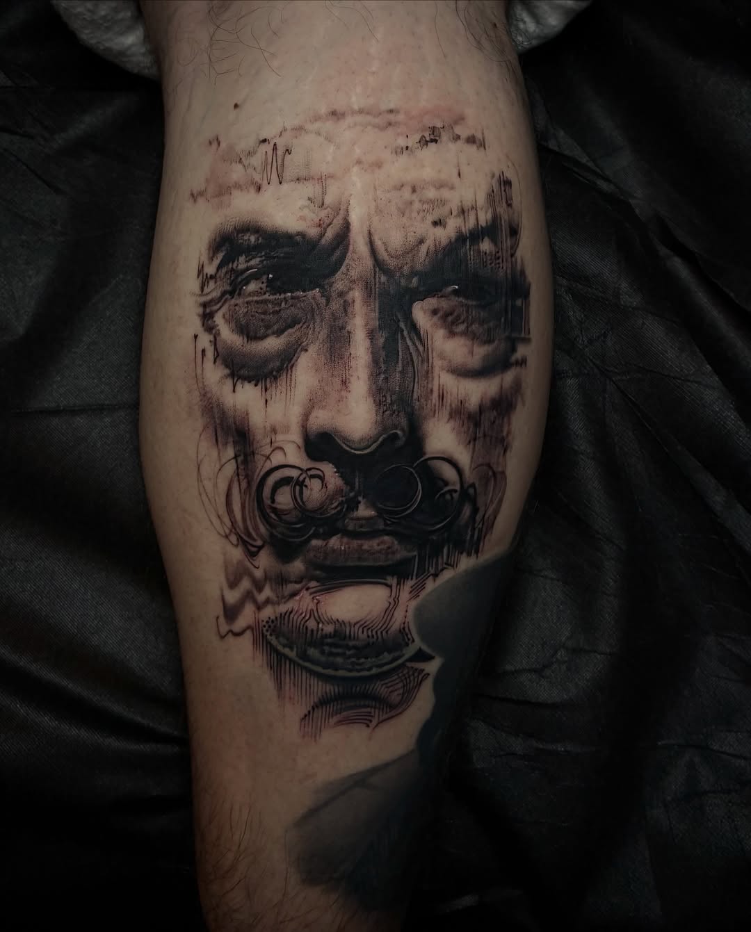 A black and grey portrait tattoo on a calf featuring a face with closed eyes, ornate mustache detail, and dripping ink effects.