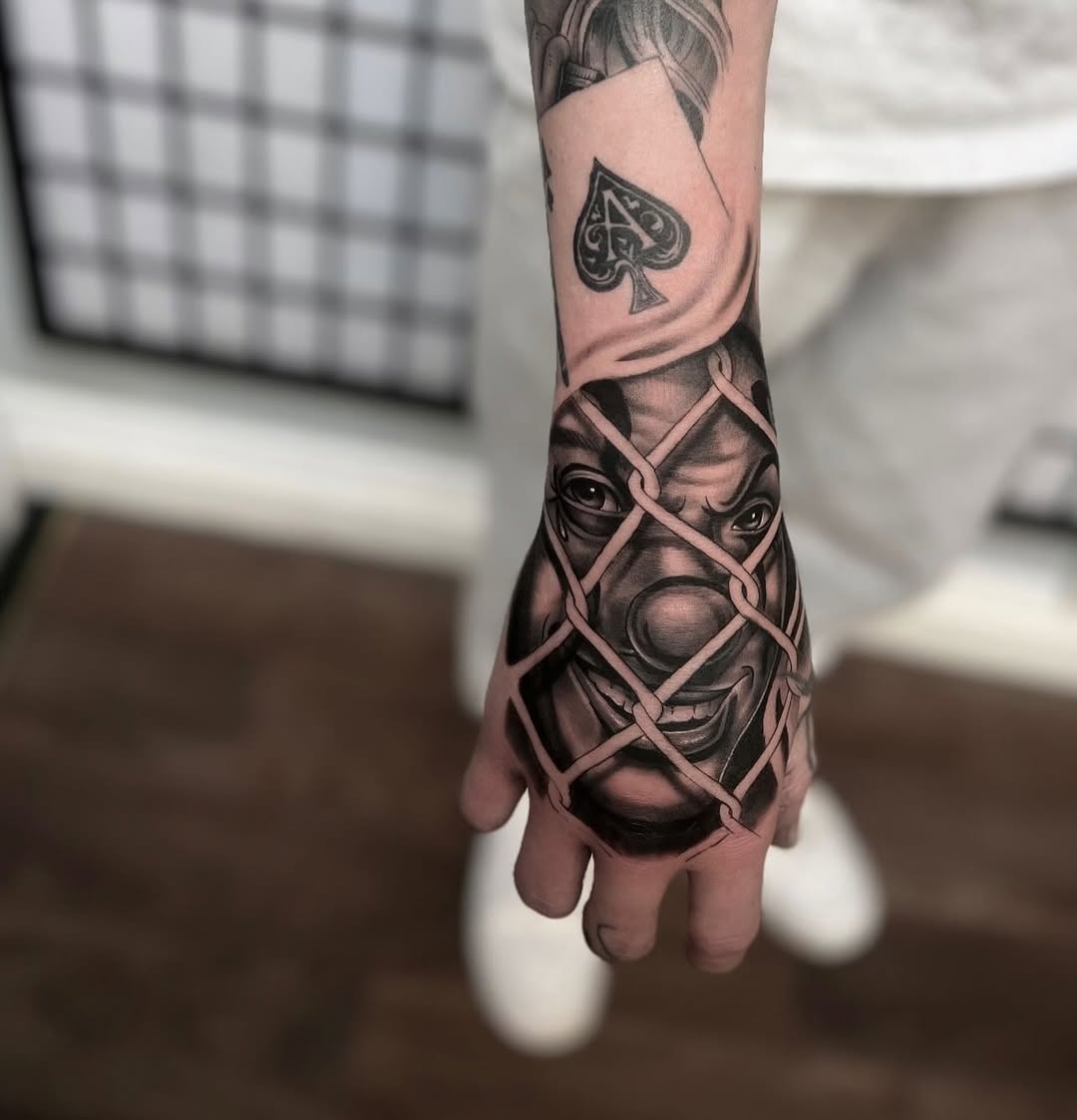 Hand tattoo featuring a demon or oni face with wrapped rope crossing the design, rendered in black and grey with realistic shading.
