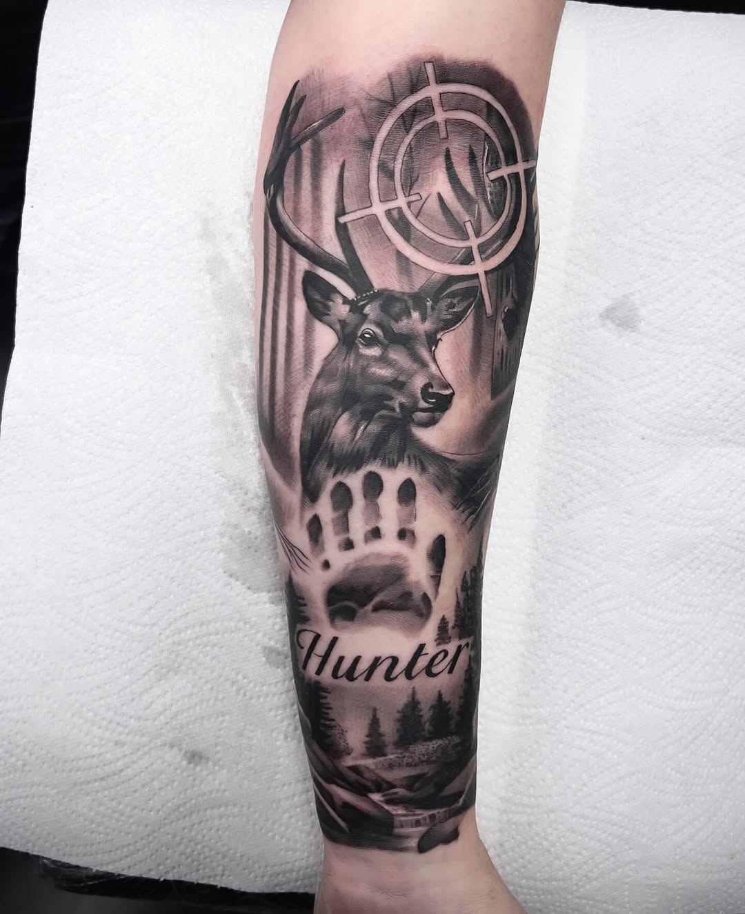 Deer head with antlers and rifle scope crosshairs on forearm, paw print below, forest landscape and 'Hunter' text, black and grey shading.