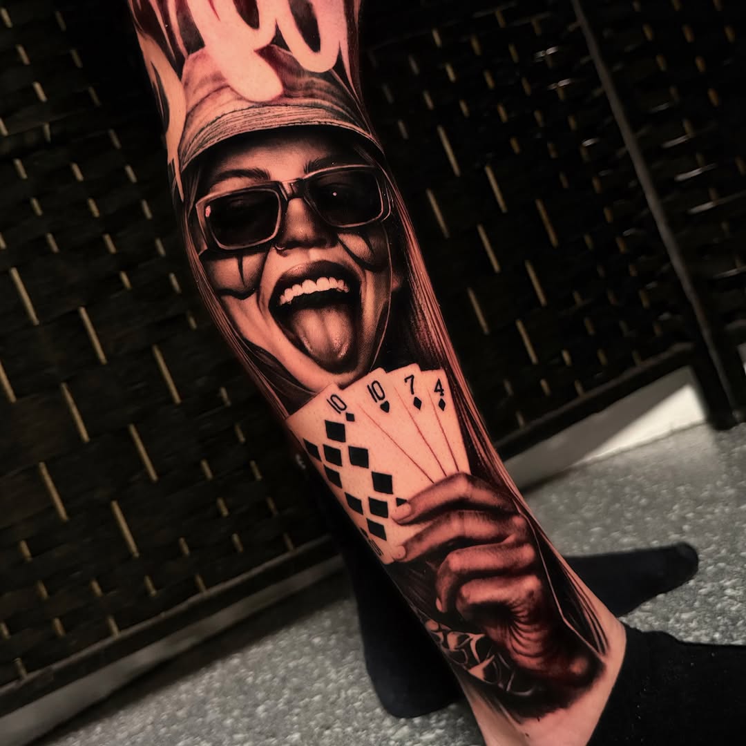 Portrait tattoo of a person with sunglasses and tongue out, holding playing cards, rendered in black and grey shading on forearm.
