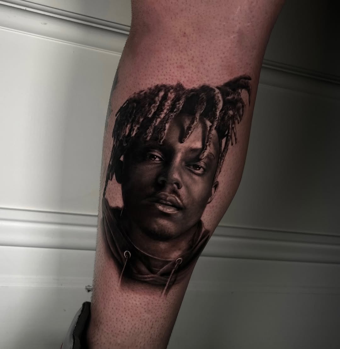 Black and grey portrait tattoo of a man's face with distinctive hair on the forearm, featuring smooth shading and realistic detail.