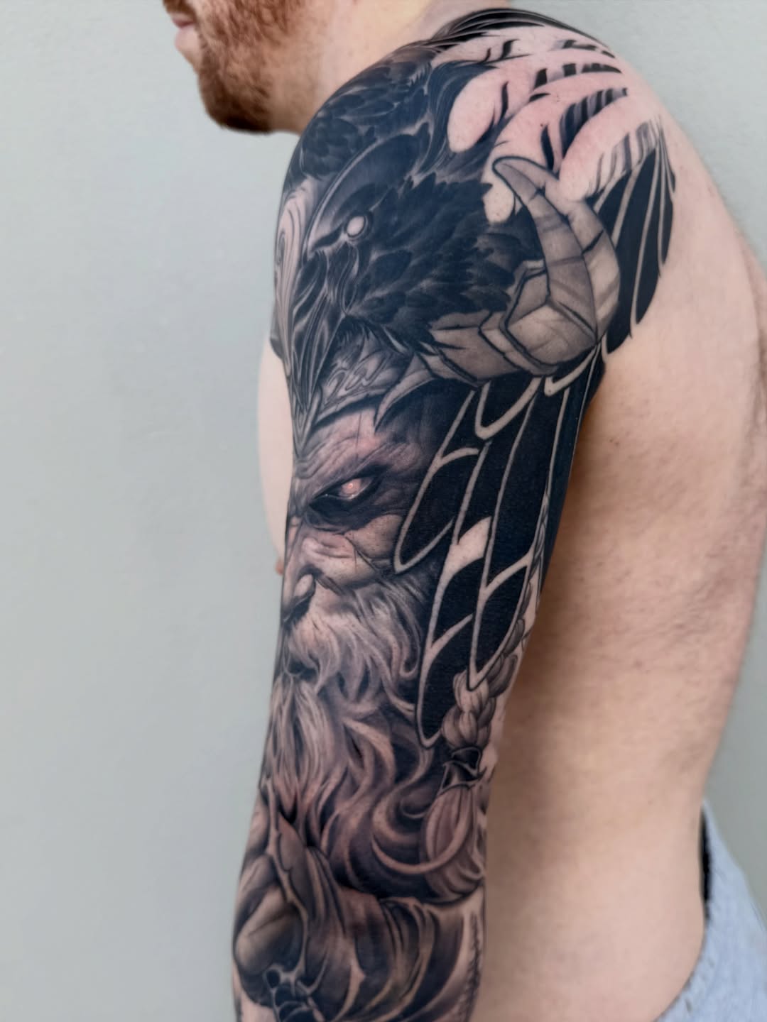 Black and grey sleeve tattoo featuring a bearded mythological face with large wings and detailed feathering on upper arm and shoulder.