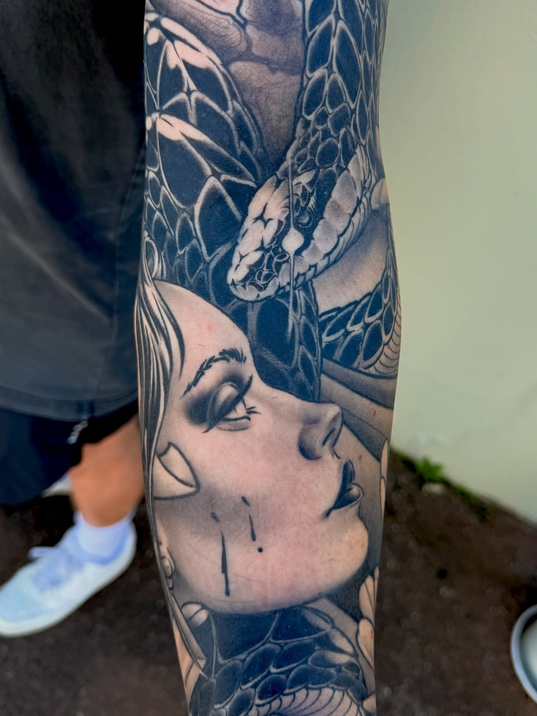 Black and grey arm sleeve tattoo featuring a woman's face with closed eyes and detailed snake and floral designs wrapping around the forearm.