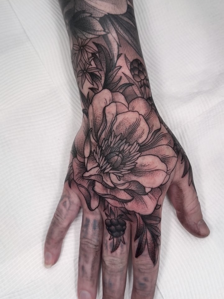 Large floral tattoo on hand and forearm featuring detailed peonies and botanical elements in black and grey with soft shading.