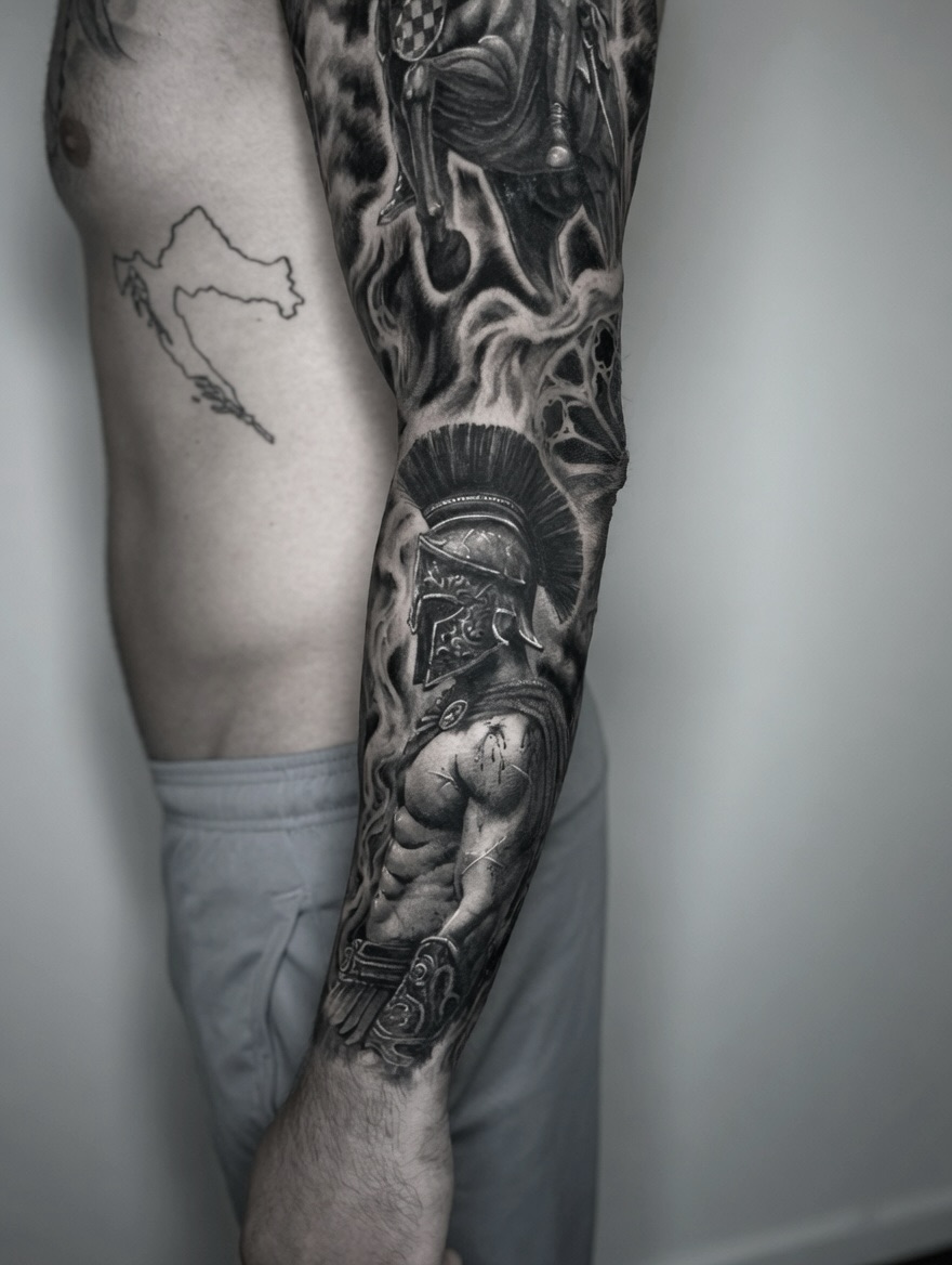 Large black and grey sleeve tattoo featuring classical warrior, demon faces, and intricate details on the arm with smooth shading.