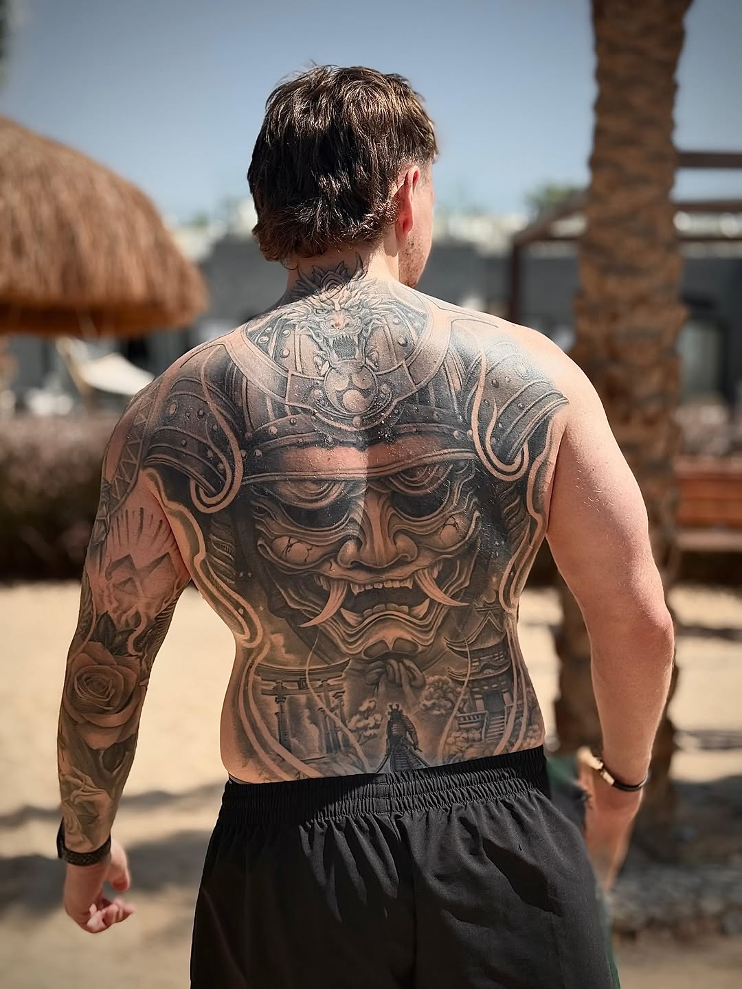 Man's back and arm feature a large samurai mask tattoo with dragon helmet, torii gates, and a rose sleeve in black and grey ink.