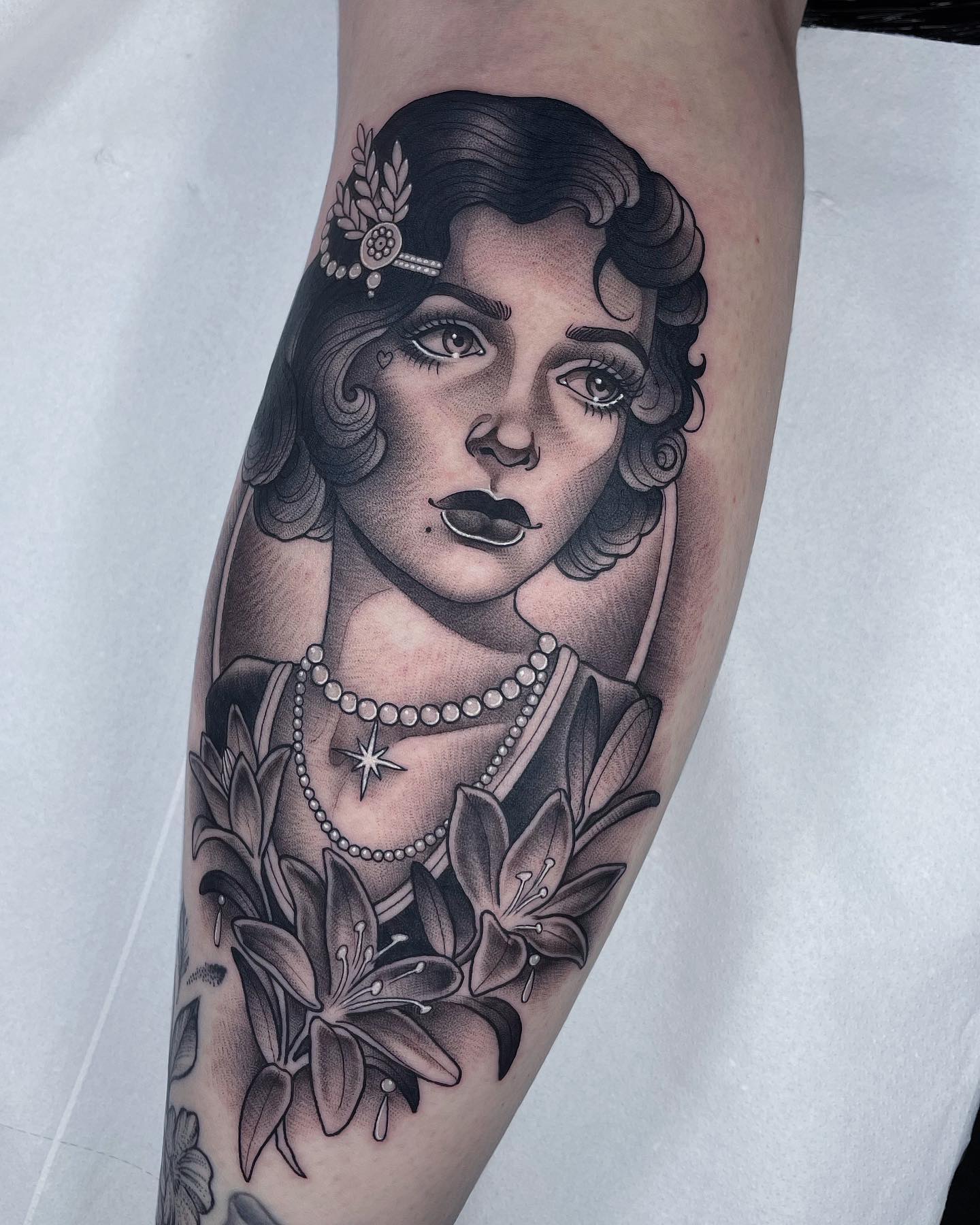 Black and grey tattoo of a vintage woman's portrait with pearl necklace and lilies on a forearm.