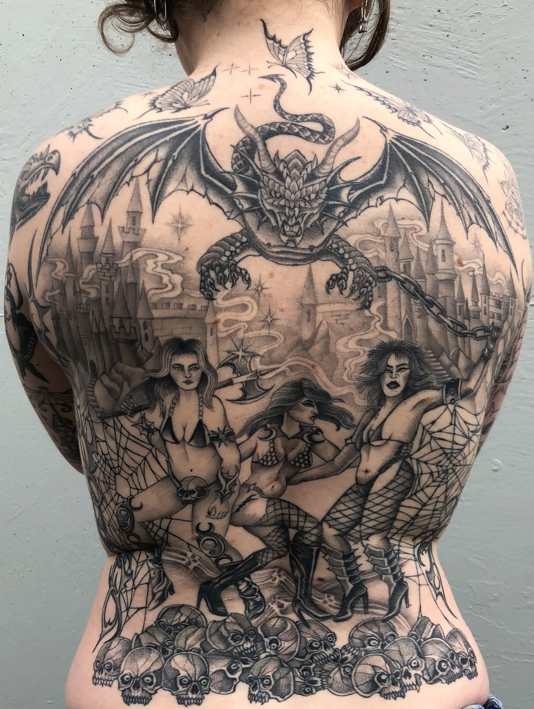Back tattoo featuring a winged demon overlooking a castle with three female figures, skulls, snakes, and butterflies in black and grey shading.