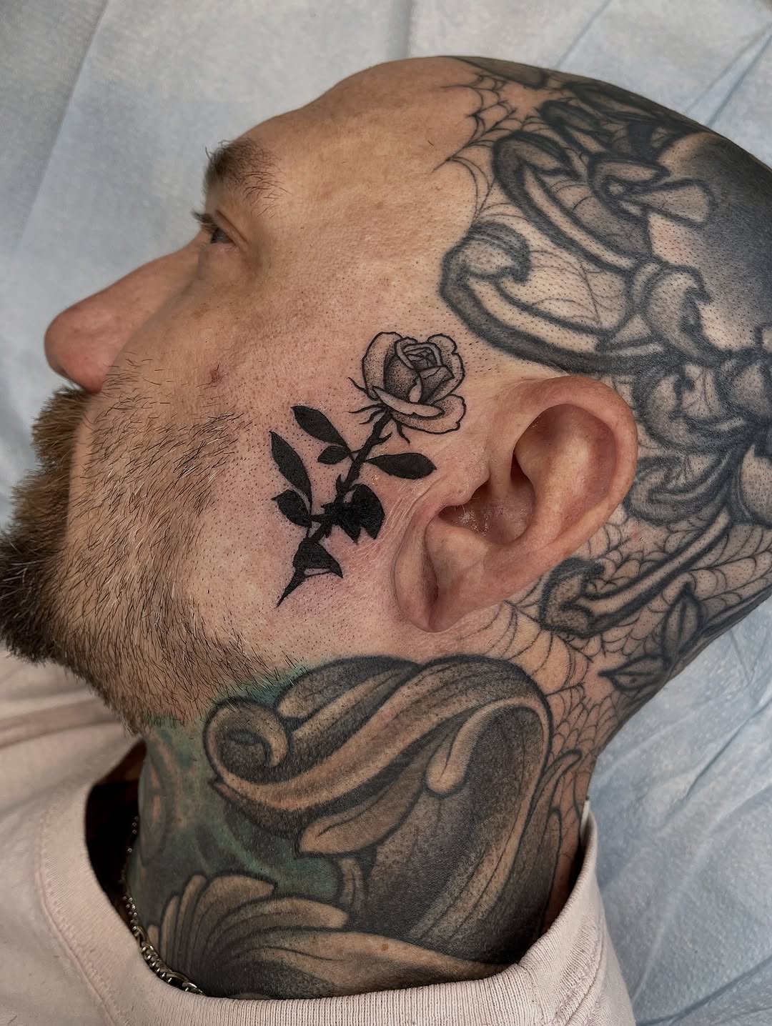 Side profile of man's head showing rose with leaves on cheek and ornate designs on scalp and neck in black and grey ink.