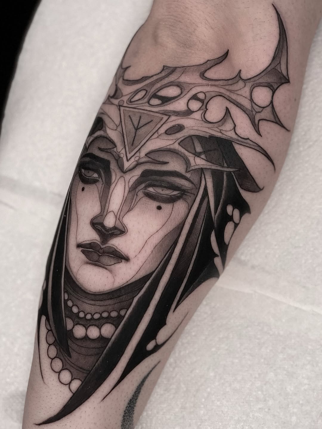 Black and grey portrait tattoo of an ornate crowned face with decorative headdress and beaded necklace on the forearm.