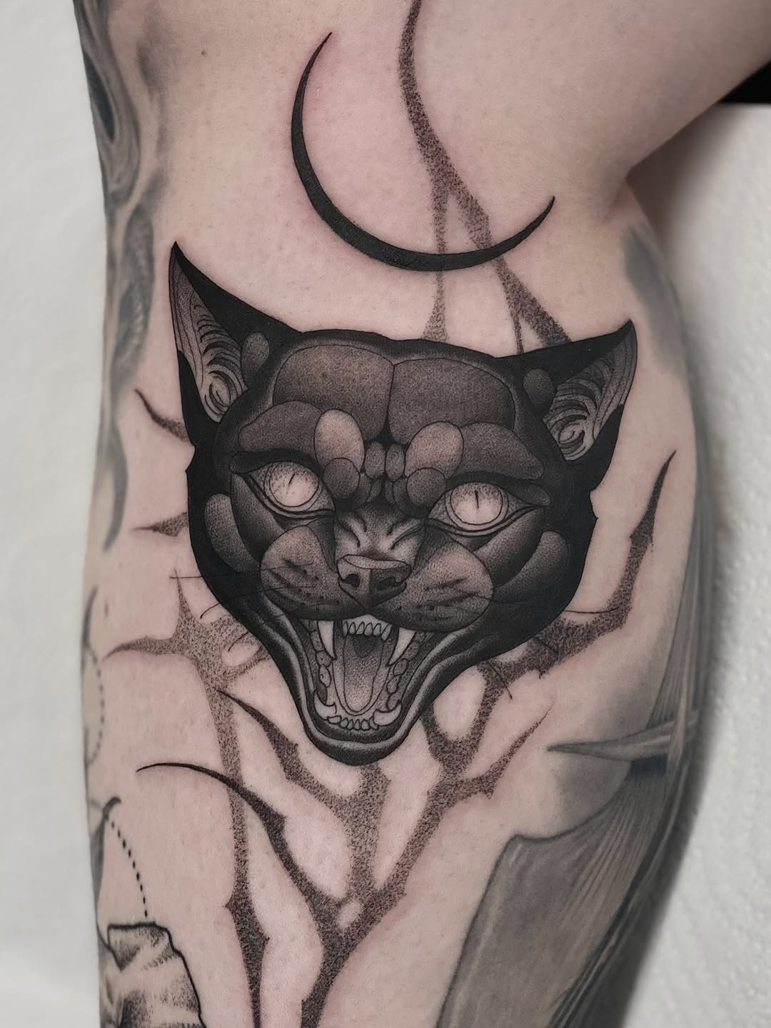 Black and grey demon cat face tattoo with crescent moons above on upper leg, featuring detailed shading and fierce expression.