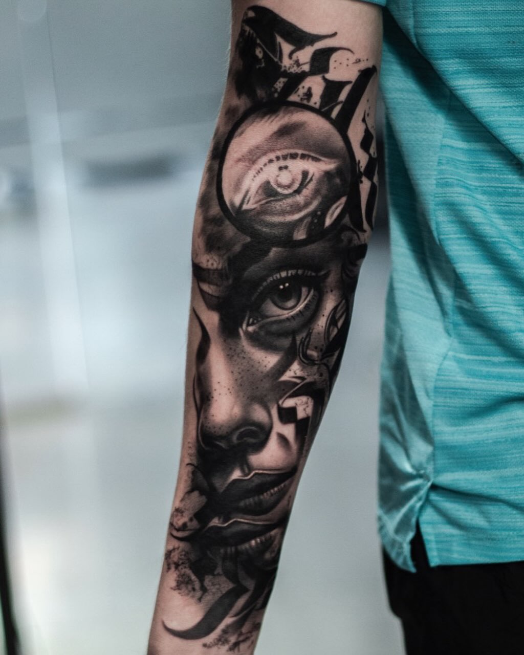 Black and grey portrait tattoo on forearm featuring stacked faces with detailed eyes and shading, worn with turquoise sleeve.