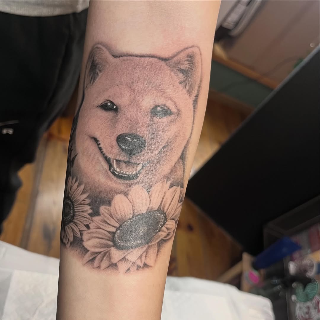 Shiba Inu dog with gentle smile surrounded by sunflowers on forearm, rendered in black and grey with soft shading.