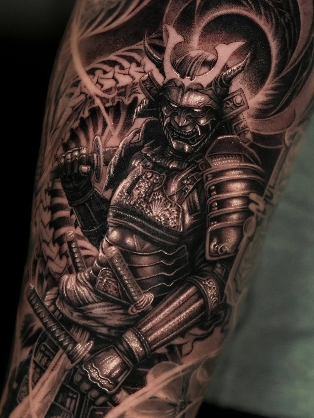 Black and grey tattoo of an ornate demon warrior in samurai armor with horns, positioned on the upper arm or shoulder.