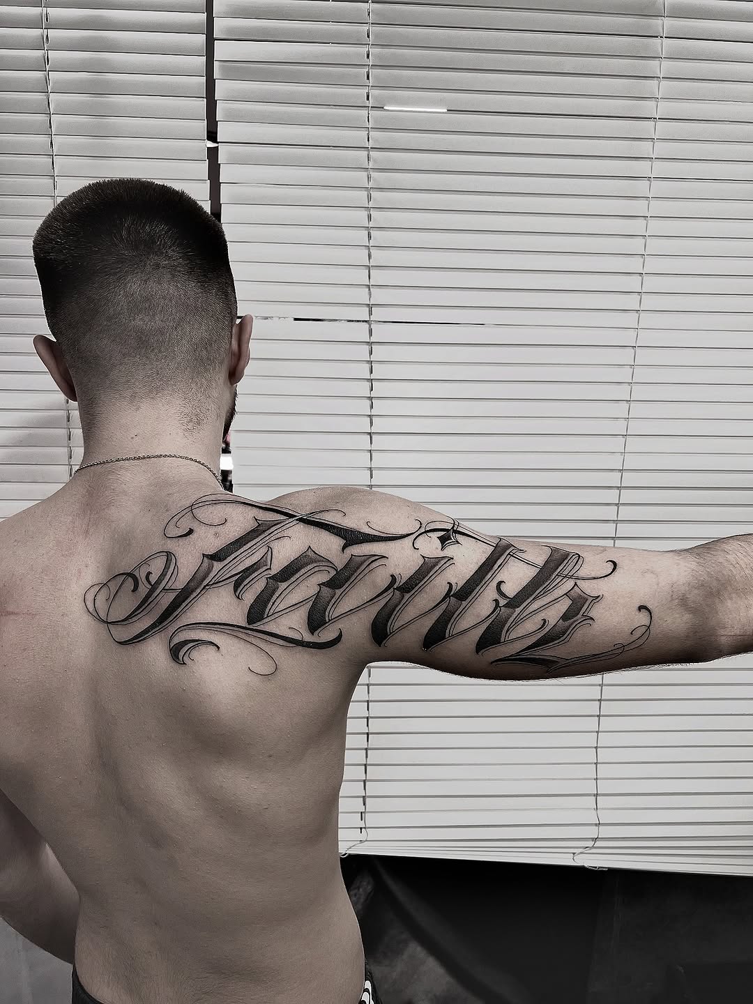 Black and grey script tattoo on upper back and shoulder blade with flowing decorative flourishes and shading.