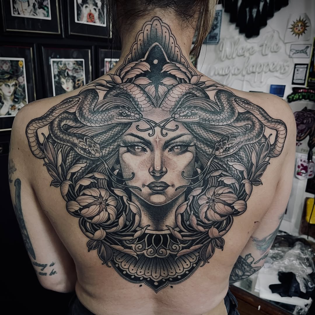 Back tattoo of a woman's face with two snakes forming a crown, surrounded by ornamental flowers and decorative elements in black and grey.