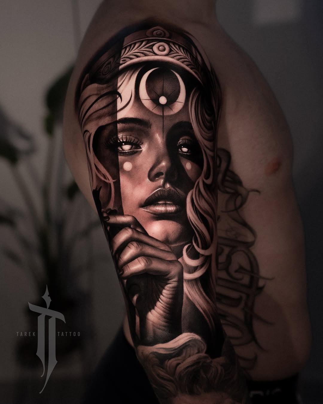 Surreal portrait tattoo on upper arm featuring a woman's face with ornate headpiece and third eye, rendered in black and grey with smooth shading.