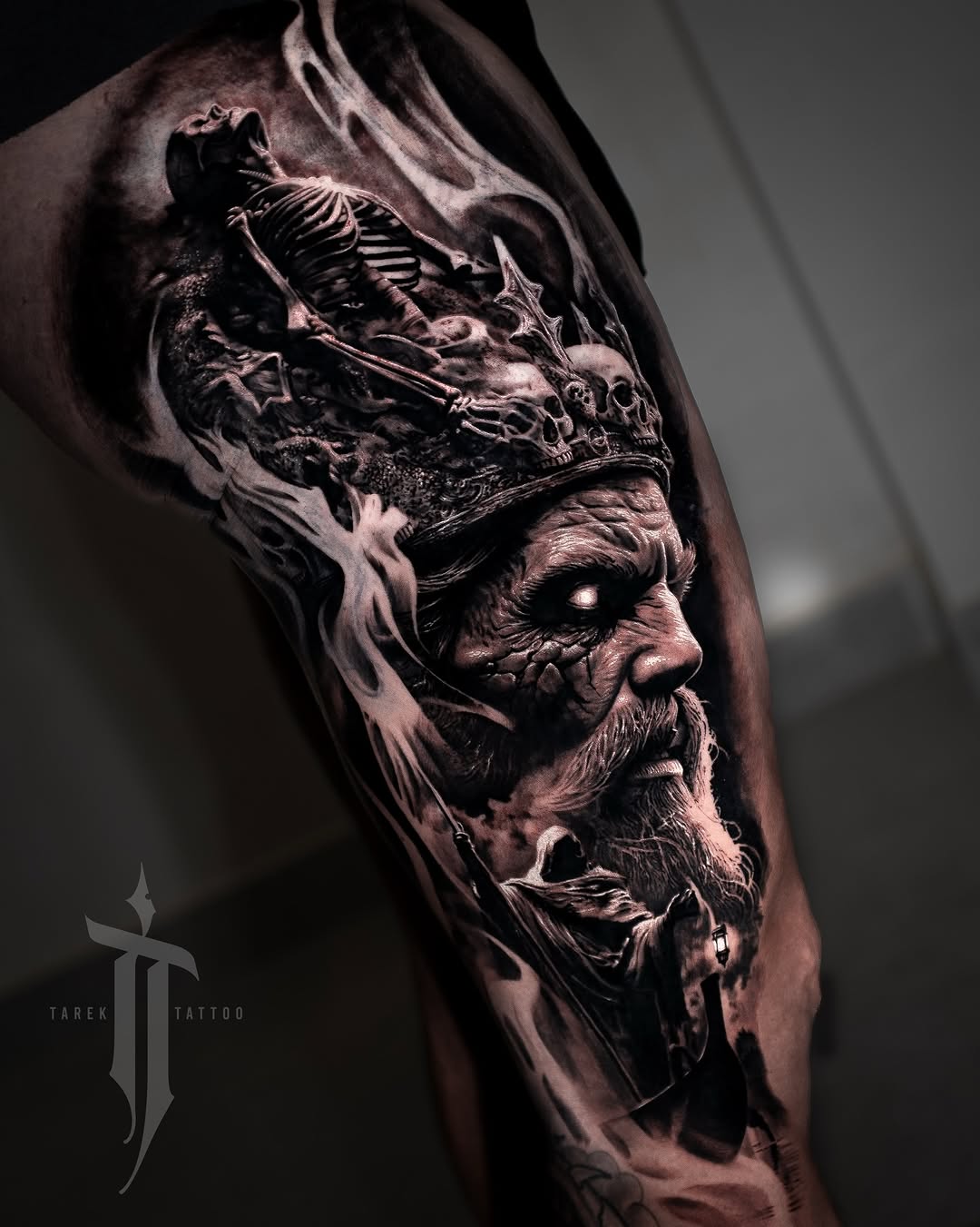 Bearded man's profile with skeletal face beneath, wearing ornate crown, rendered in black and grey realism on upper arm.