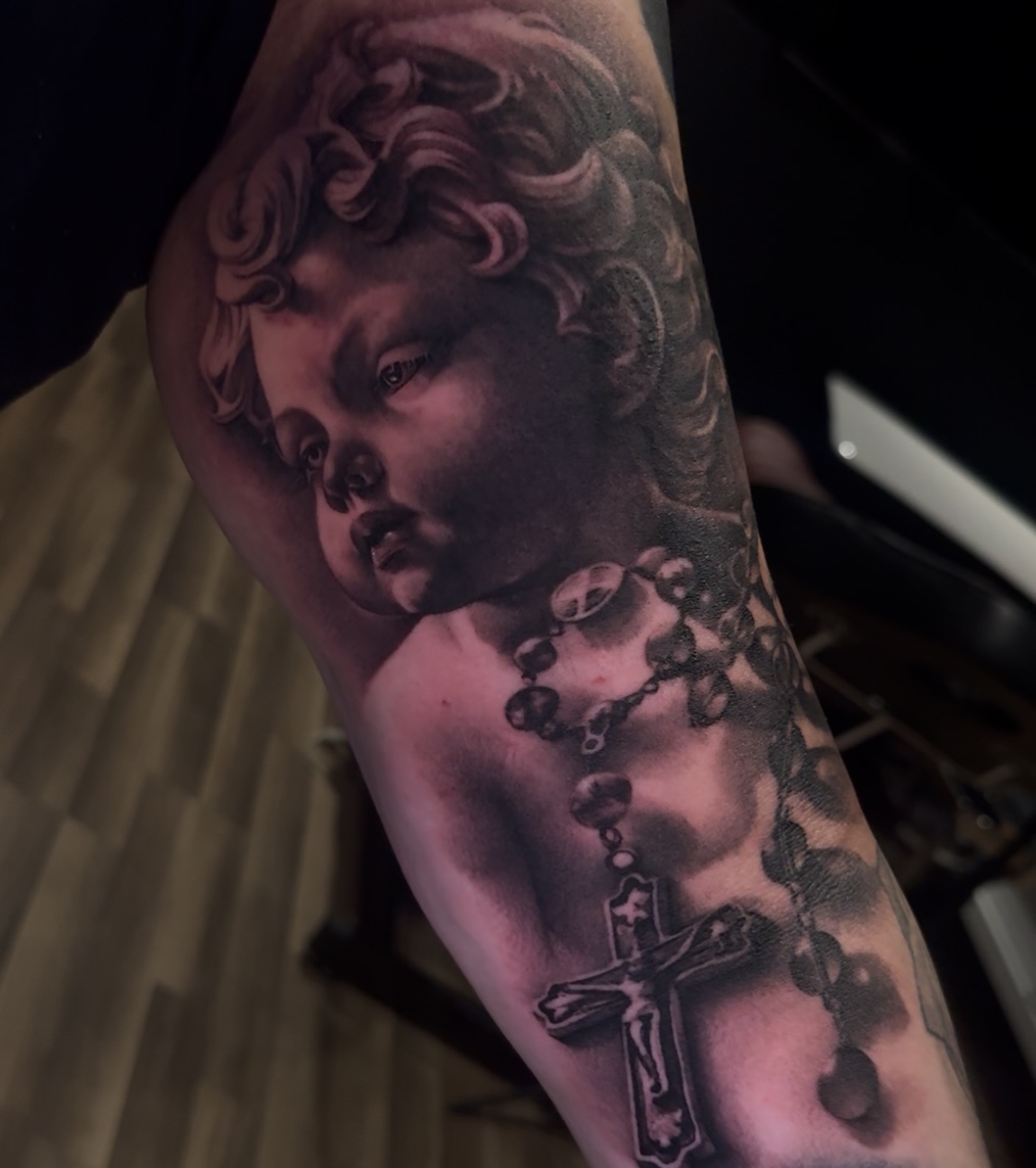 Black and grey portrait tattoo of a cherubic child's face with curled hair and a cross with rosary beads on the arm.