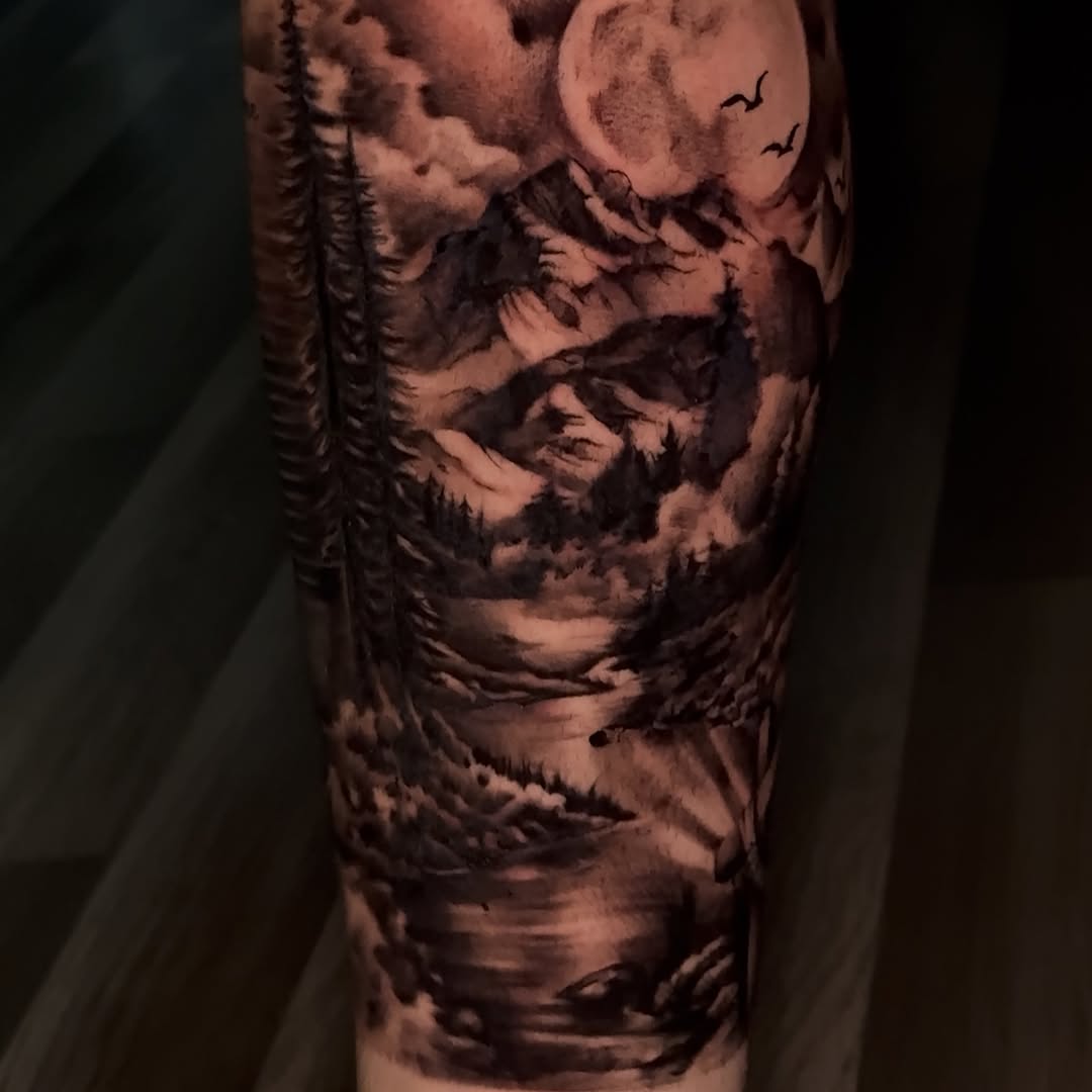 Black and grey landscape tattoo on upper arm featuring mountains, forest trees, moon, and flying birds with realistic shading.