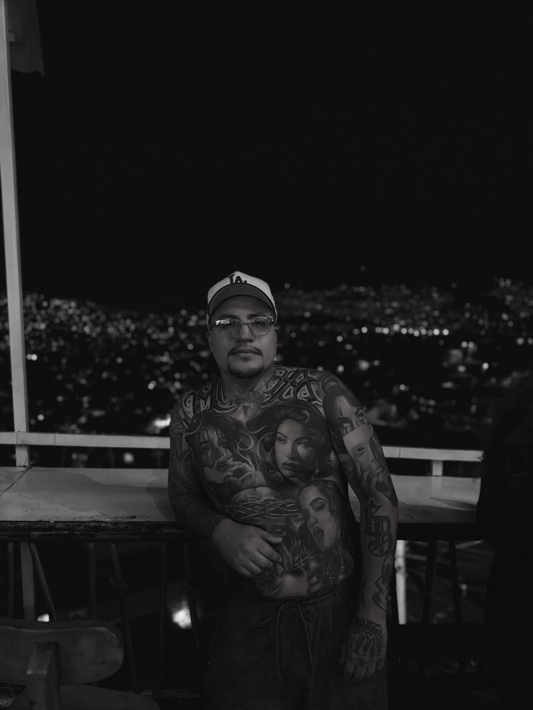 Man with extensive black and grey portrait tattoos covering his chest and arms, featuring multiple faces with detailed shading, photographed at night with city lights behind him.