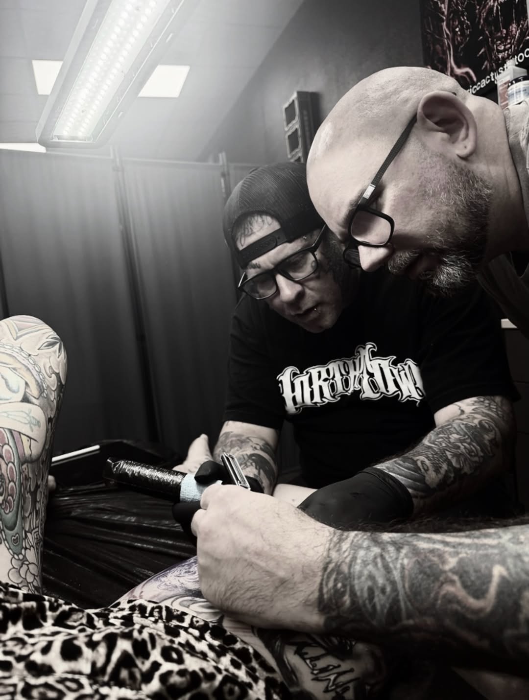 Tattoo artist applying black and grey ink to client's arm with elaborate shaded designs visible on both figures' skin.